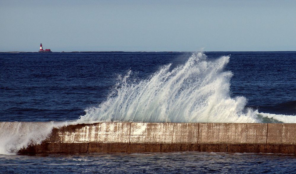 Photograph of a wave cresting over a seawall with a lighthouse in the distant background..jpg