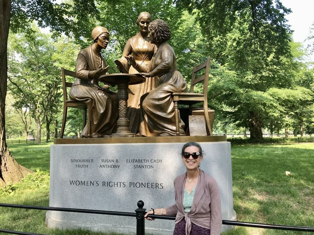Photo of the author standing in front of the Women’s Rights Pioneers Monument. The sun shines on the statues of the pioneers..jpg