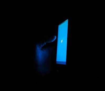 in total darkness a hand holds up a smartphone displaying a blue screen with the twitter logo.jpg