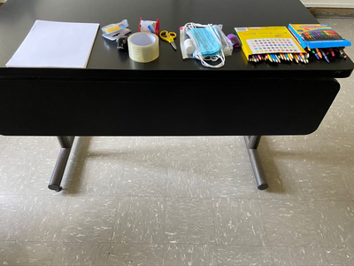 Art supplies placed on a classroom table, including paper, tape, scissors, surgical masks, gauze, stapler, colored pencils, and crayons..png