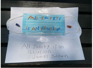 Blue surgical mask with quote 'All safety is an illusion' in blue and orange crayon, James Baldwin's name, and broken blue and orange crayon tips on ear loops..png