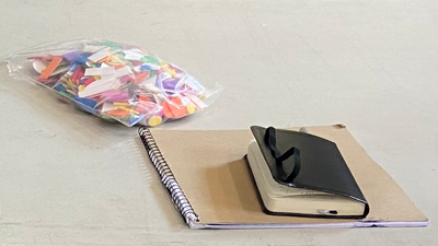 A plastic bag of multicolored paper scraps rests on a table next to a sketch book with a small black journal on top. Photo by Susan Bernstein, October 19 2023
