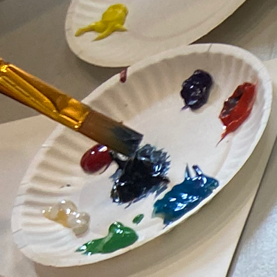 Paintbrush hovers above paper plate with acrylic pain in rainbow colors. Photo by Susan Bernstein, October 19 2023