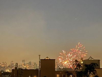 Fireworks exploding over apartment buildings near the East River with the NYC skyline in the background. Photo by Susan Bernstein, July 4, 2023