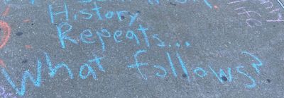 Chalk art on a campus sidewalk. Blue chalk on pavement: reads History Repeats…What follows?