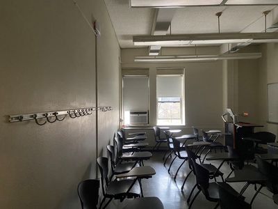 An empty classroom. Coat hooks on the back wall, 3 rows of desks behind a computer console. Two windows, one with an air conditioner, the other partially opened with bare trees in the background. The window shades let in light. Photo by Susan Bernstein December 7, 2023.