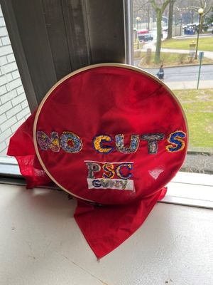 Red banner embroidered with NO CUTS PSC CUNY  placed on a classroom window ledge.  The window overlooks the boulevard in front of campus.