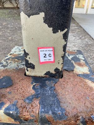A sticker that reads Give Us Your Two Cents on a rusted pole outside a classroom building.jpg